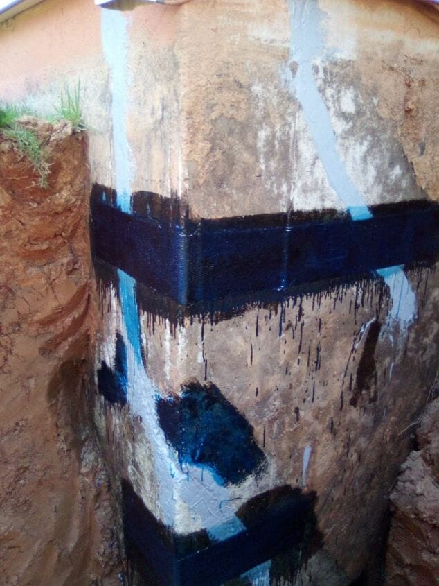 Concrete foundation wall with visible waterproofing membrane and sealant applied at seams, highlighting areas commonly targeted for foundation crack repair, partially surrounded by excavated soil.